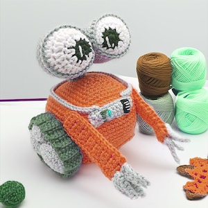 Puede incluir: Un juguete de robot de ganchillo naranja hecho a mano con grandes ojos blancos y grises. El robot tiene un borde, brazos y una rueda grises y blancos. Varios carretes de hilo en marrón, gris y verde están en el fondo. También se ven una pequeña bola verde y una hoja de ganchillo.