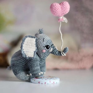 May include: A crocheted gray elephant with large black eyes and pink cheeks holds a pink heart-shaped balloon. The elephant has white-lined ears and sits on a small, round, patterned base. The balloon is attached with white string.