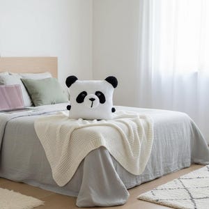 Puede incluir: Una habitación con una cama con ropa de cama gris, una manta de punto crema y un cojín con forma de panda. Hay un portátil en la cama. La habitación tiene una cabecera de madera clara, una alfombra pequeña y una ventana con cortinas transparentes.