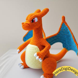 May include: Crocheted orange dragon with blue wings and a cream-colored belly. The handmade plush toy has white accents on its feet and a detailed face with red eyes. The dragon is sitting on a white surface.