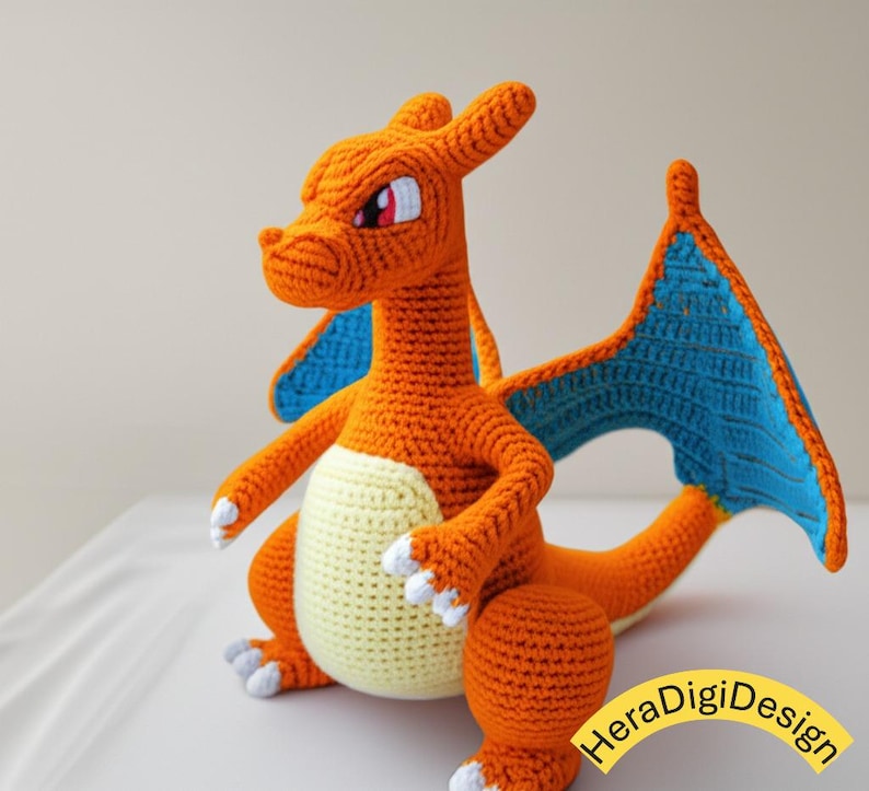May include: Crocheted orange dragon with blue wings and a cream-colored belly. The handmade plush toy has white accents on its feet and a detailed face with red eyes. The dragon is sitting on a white surface.