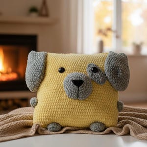 Puede incluir: Un cojín de perro cuadrado hecho a mano con hilo amarillo suave y detalles grises. El cojín presenta una cara de perro con orejas, nariz y parches oculares grises, y pequeños pies grises. Está sobre una manta beige.