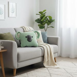 May include: A light gray sofa with a green koala-shaped pillow, a green textured pillow, and a blue and white patterned pillow. A cream-colored blanket is draped over the sofa. Two framed botanical prints hang on the wall.