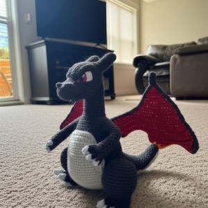 Könnte beinhalten: Ein handgehäkelter Drache in Dunkelgrau mit weißem Bauch und roten Flügeln. Der Drache hat rote Augen und weiße Akzente an Füßen und Krallen. Der Drache sitzt auf einem Teppichboden.