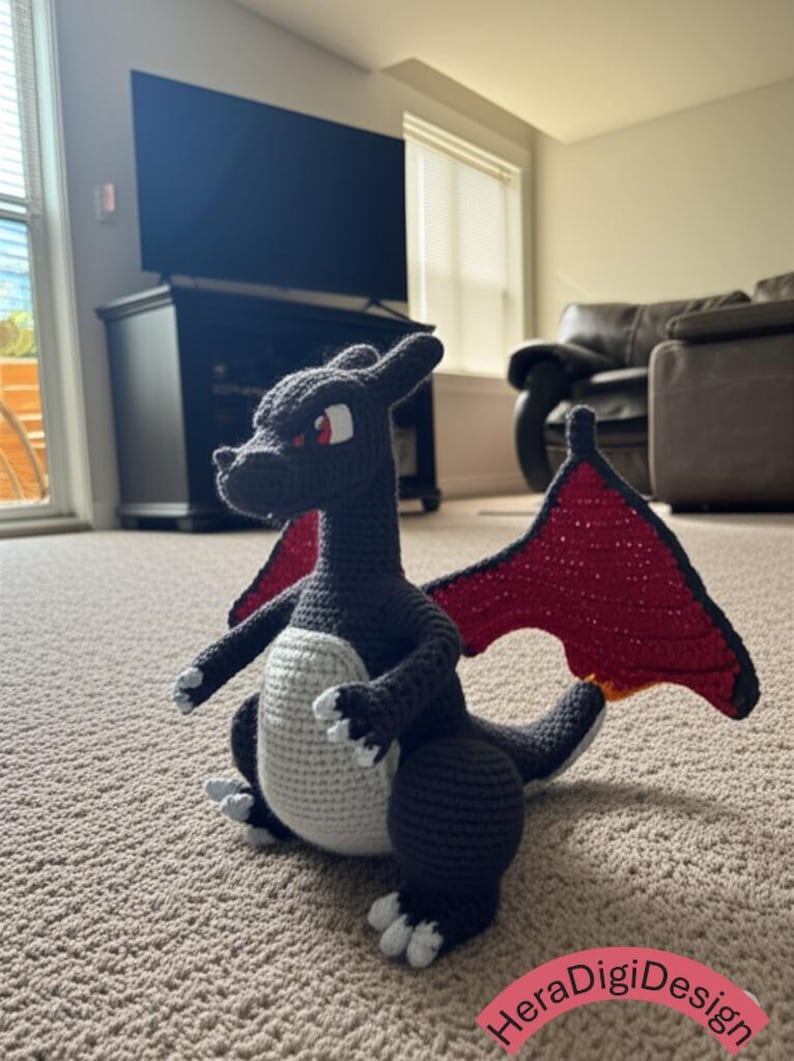 May include: A handmade crochet dragon in dark gray, with a white belly and red wings. The dragon has red eyes and white accents on its feet and claws. The dragon is sitting on a carpeted floor.