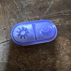 Puede incluir: Un recipiente de plástico azul y blanco con un diseño de sol y luna. El recipiente está dividido en dos secciones, una con un sol y otra con una luna creciente. El recipiente es rectangular con bordes redondeados.