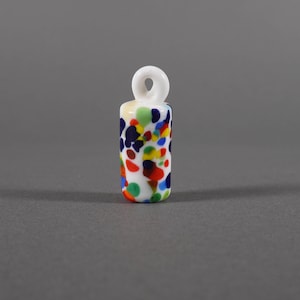 Może przedstawiać: Mały, biały szklany wisiorek z pętelką na górze. Cylindryczny korpus jest ozdobiony kolorowymi plamkami w kolorze czerwonym, niebieskim, żółtym i zielonym. Wisiorek jest prawdopodobnie przeznaczony do tworzenia biżuterii lub rękodzieła.