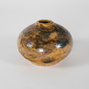 Op de afbeelding: Een kleine, ronde keramische vaas met een smalle opening. De vaas heeft een gevlekt oppervlak met tinten bruin, beige en blauw. Het keramische stuk heeft een rustieke, handgemaakte uitstraling.
