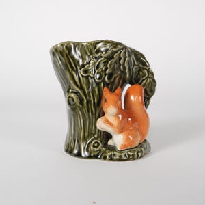 May include: A ceramic figurine of a squirrel next to a tree trunk. The tree trunk is dark green with detailed bark and leaves. The squirrel is orange and white, with a fluffy tail. The figurine is likely a decorative item.