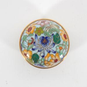 Puede incluir: Una caja de baratijas redonda y decorativa con un fondo azul claro y un diseño floral. Las flores son en tonos de azul, amarillo, naranja, verde y rosa. La caja tiene un borde dorado.