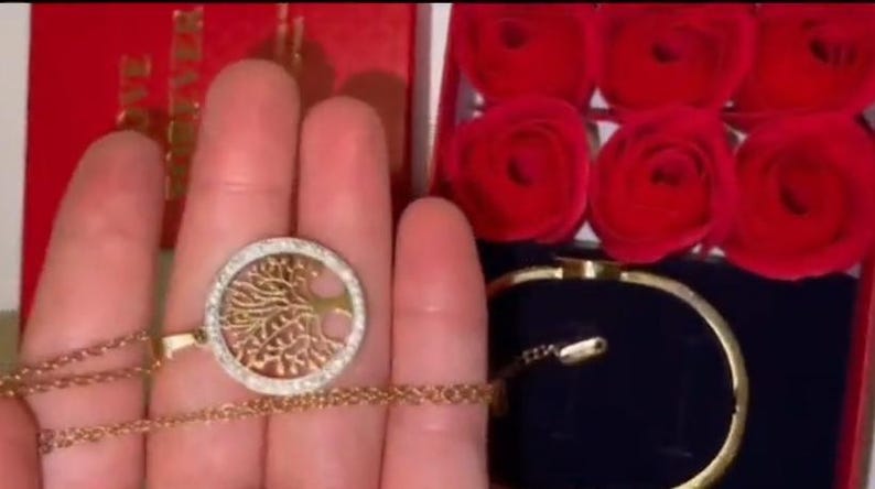 May include: A gold necklace with a tree of life pendant and a matching gold bracelet. The necklace and bracelet are in a red box with a white background. The box is filled with red roses.