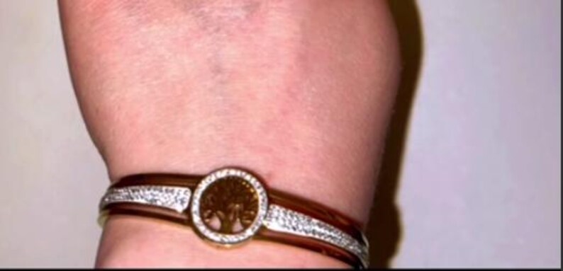 May include: A gold bangle bracelet with a clear crystal design and a round, gold charm featuring a tree design.