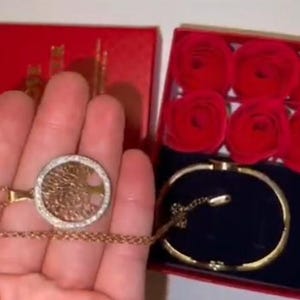May include: A gold necklace with a tree of life pendant and a gold bracelet in a red box with six red roses.