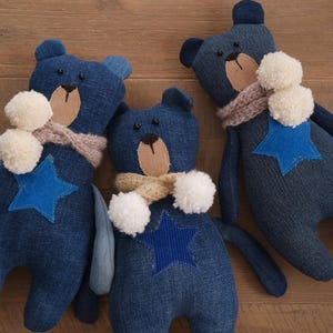 May include: Three handmade denim bear plush toys. Each bear has a light brown muzzle, a blue star applique, a cream-colored pom-pom, and a scarf. The bears are arranged on a wooden surface.
