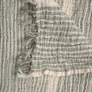 May include: A close-up shot of a textured blanket with green and off-white stripes. The blanket has a crinkled texture and a fringed edge. The vertical stripes alternate in colour, creating a soft and inviting appearance.