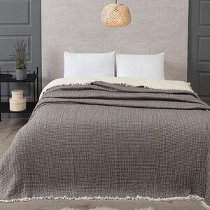 Op de afbeelding: Een grijze sprei met een textuurpatroon bedekt een bed met witte kussens. De sprei heeft een gefranjerde rand. Boven het bed hangt een geweven hanglamp. Er ligt een klein vloerkleed op de grond.