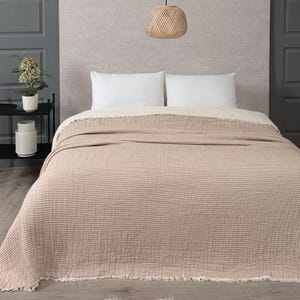 May include: A bed is made with white pillows and a beige blanket with a textured grid pattern. The blanket has a fringed edge. A woven pendant light hangs above the bed. A small black side table holds a plant and a decorative vase.