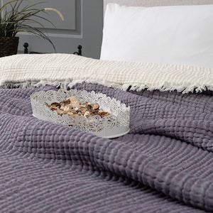 Könnte beinhalten: Ein weißes, ovales Dekotablett mit filigranem Lochmuster, gefüllt mit getrocknetem Potpourri, liegt auf einer strukturierten, lila Bettdecke. Eine weiße Decke mit Fransen und ein Kissen sind im Hintergrund zu sehen.