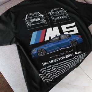 May include: Black t-shirt featuring a blue BMW M5 sedan graphic with white outlines and text. The design includes the BMW logo and the words "THE MOST POWERFUL BMW." The shirt also has technical drawings of the car.