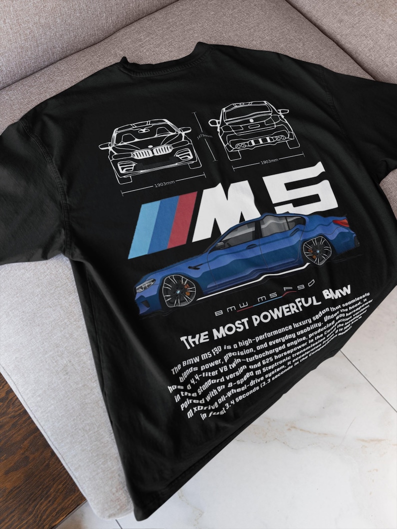 May include: Black t-shirt featuring a blue BMW M5 sedan graphic with white outlines and text. The design includes the BMW logo and the words "THE MOST POWERFUL BMW." The shirt also has technical drawings of the car.