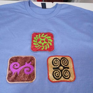 Könnte beinhalten: Hellblaues T-Shirt mit drei gestickten Aufnähern. Die quadratischen Aufnäher mit glitzernden Rändern zeigen Designs in Rot, Grün, Lila und Gold. Die Designs umfassen einen Sonnenstrahl, eine Wirbel und eine Reihe von Spiralen.