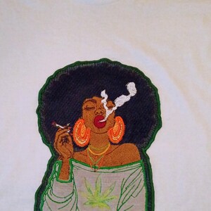 Puede incluir: Una camiseta blanca con un gran parche bordado de una persona con peinado afro oscuro, que lleva grandes pendientes de aro naranjas y una blusa gris claro de hombros descubiertos con un gráfico de hoja de cannabis verde, sosteniendo un cigarrillo.