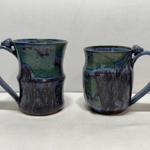 May include: Two ceramic mugs featuring a mottled glaze in shades of deep blue, dark purple, and forest green. Each mug has a curved handle with a small, rounded thumb rest at the top. The mugs have a cylindrical shape with a slightly flared rim.