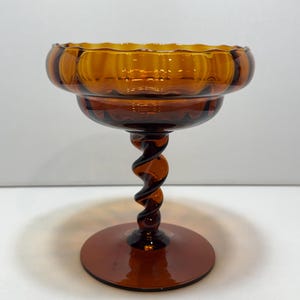 May include: Amber-colored glass pedestal bowl with a spiral stem and a scalloped rim. The bowl has a wide, round base and a decorative, textured design. The glass has a warm, inviting hue, perfect for displaying small items or serving treats.