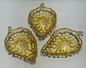 May include: Three amber-coloured glass leaf-shaped dishes. Each dish has a textured surface with a pattern of small circles and a central fan design. The dishes have a small handle at the top.