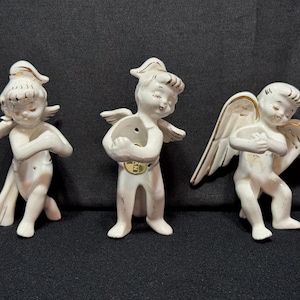 Peut inclure: Trois figurines de chérubins en céramique blanche avec des détails dorés. Chaque chérubin a les yeux fermés, des ailes et une pose unique. L'un est agenouillé, un autre tient un objet rond, et le troisième a les mains jointes.
