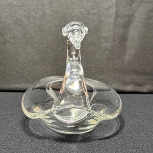 May include: Clear glass swan-shaped dish. The swan has a long neck and a flared base, with a circular dish forming the body. The glass is transparent, allowing light to pass through. The dish is likely for decorative purposes.