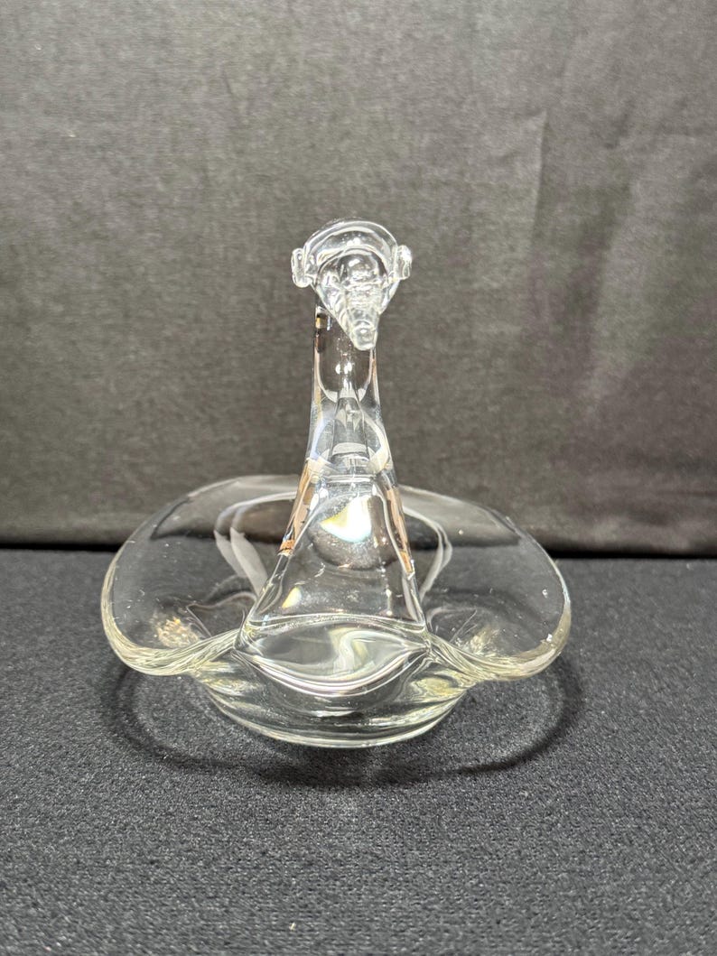 May include: Clear glass swan-shaped dish. The swan has a long neck and a flared base, with a circular dish forming the body. The glass is transparent, allowing light to pass through. The dish is likely for decorative purposes.