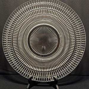 Può includere: Un piatto di vetro trasparente con un design circolare. Il piatto presenta anelli concentrici di linee verticali e un motivo centrale a forma di raggiera. Il piatto è esposto su un supporto nero su sfondo scuro.