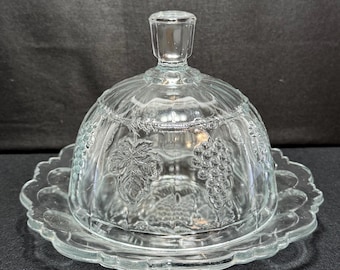 May include: A clear glass butter dish with a domed lid and a scalloped edge base. The lid has a handle and embossed grape and leaf designs. The dish is designed to hold butter and keep it fresh.