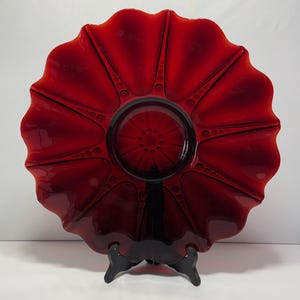 May include: A ruby red glass plate with a scalloped edge and a central design. The plate is supported by a black stand, showcasing its intricate details and vibrant color. The plate's design features radial lines and circular accents.