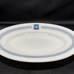 May include: White oval serving platter with a blue Greek key border design. The plate has a small blue emblem on the top edge. The plate is made of a glossy material and is set against a dark background.