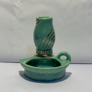 May include: A teal ceramic candle holder featuring a rounded base with a textured lattice pattern and a small handle. The central cylindrical candle cup is decorated with gold-colored diagonal stripes and a gold-colored band at the base.