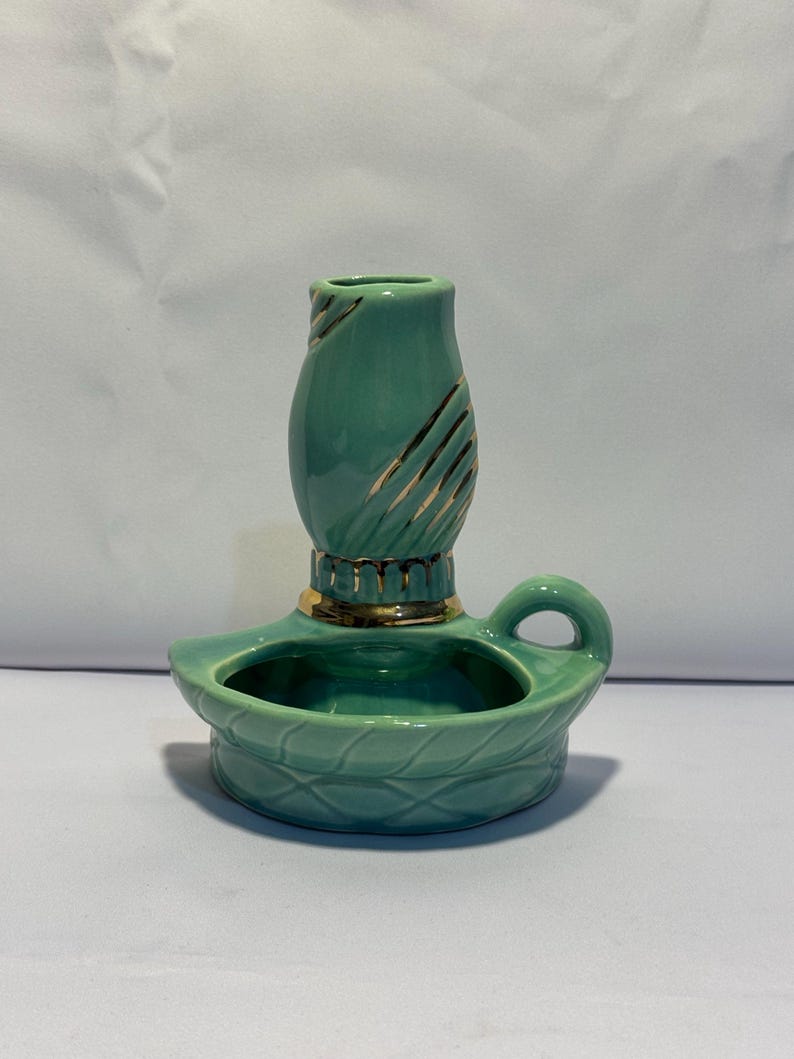 May include: A teal ceramic candle holder featuring a rounded base with a textured lattice pattern and a small handle. The central cylindrical candle cup is decorated with gold-colored diagonal stripes and a gold-colored band at the base.