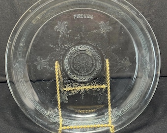 May include: Clear glass plate with decorative floral and scrollwork design. The plate has the text "FIRE-KING" embossed on the rim. The plate is displayed on a gold-colored metal stand.