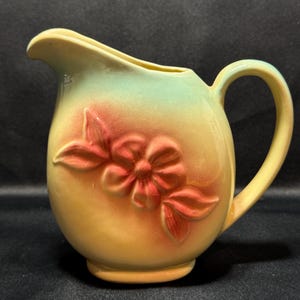 Puede incluir: Una jarra de cerámica con cuerpo redondeado y asa. La jarra es principalmente amarilla, con un degradado de verde en la parte superior y un diseño floral en relieve en tonos rojos y rosas. El pico es curvo.