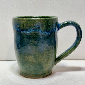 Puede incluir: Una taza de cerámica hecha a mano con un cuerpo redondeado y un asa curva. La taza presenta un esmalte verde y azul, creando un efecto veteado. La taza está diseñada para bebidas calientes.
