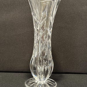 Könnte beinhalten: Eine klare Glasvase mit einem ausgestellten, gewellten Rand und einem rautenförmigen Körper. Die Vase hat einen abgerundeten Boden und einen kreisförmigen Fuß. Das Glas ist transparent und eignet sich zur Präsentation von Blumen. Die Vase ist ca. 20 cm hoch.