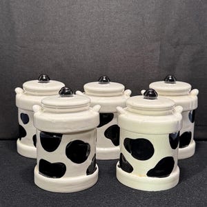 May include: Five ceramic canisters with lids. The canisters are white with black cow spots. Each canister has a small black knob on the lid and two small handles. The canisters are arranged on a dark surface.