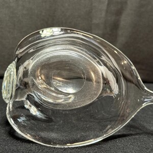May include: A clear glass serving dish with a unique, asymmetrical shape. The dish has a rounded bowl with a pointed spout on one side and a small, decorative handle on the other. The glass is transparent, allowing light to pass through.