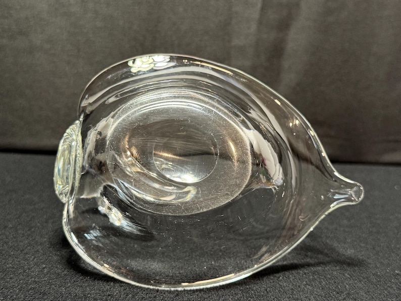 May include: A clear glass serving dish with a unique, asymmetrical shape. The dish has a rounded bowl with a pointed spout on one side and a small, decorative handle on the other. The glass is transparent, allowing light to pass through.