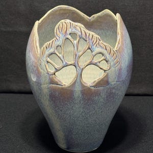 May include: A handcrafted ceramic vase with a unique tree-like cutout design. The vase has a blue and purple iridescent glaze, with a textured surface. The rim has a scalloped edge. The vase is a decorative item.