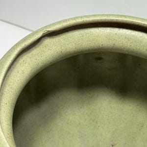 May include: Close-up of a light green ceramic bowl with a slightly rough texture. The rim of the bowl has a small chip. The interior of the bowl is smooth and the color is consistent throughout.