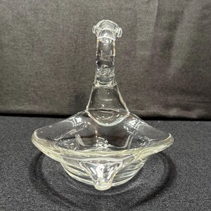 May include: Clear glass swan-shaped dish with a flared base and a long, curved neck. The dish has a shallow, square-shaped bowl. The glass is transparent, allowing light to pass through, and the design is simple and elegant.
