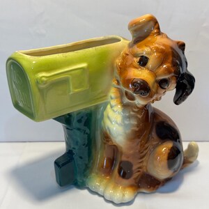 May include: A ceramic dog planter with a green mailbox. The dog is brown, tan, and black with a white chest and paws. The mailbox is light green with the words "U.S. MAIL" on the side. The planter is a vintage-style decorative item.