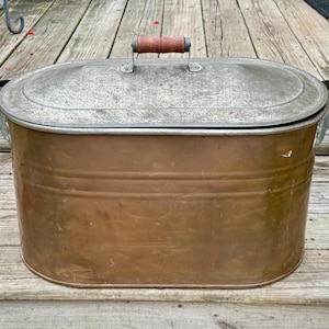 May include: An oval copper wash boiler with a removable galvanized metal lid. The container features two side handles and a central lid handle, all crafted from brown wood. The copper body has horizontal ridges and a weathered metallic finish.